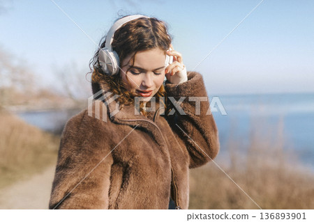 Young woman listening to music with headphones by the sea on a sunny day. Relaxed mood, closed eyes, enjoying sound and nature, modern lifestyle and calm outdoor moment. 136893901