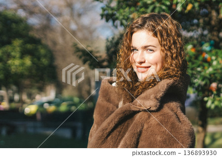 Portrait of young woman outdoors in natural light. Curly hair, warm sunlight, green background, calm expression, lifestyle beauty concept in nature. Portrait of young woman outdoors in natural light. Curly hair, warm sunlight, green background, calm expression, lifestyle beauty concept in nature. 136893930