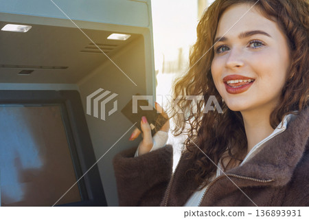 Smiling young woman using ATM outdoors, holding bank card and withdrawing cash. Modern lifestyle, personal finance, banking service, urban environment, daylight, positive emotion. Smiling young woman using ATM outdoors, holding bank card and withdrawing cash. Modern lifestyle, personal finance, banking service, urban environment, daylight, positive emotion. 136893931