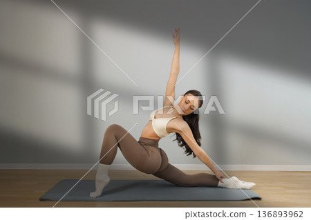 Flexible young woman practicing yoga stretch indoors. Female performing deep lunge side stretch pose on yoga mat, strength, balance and flexibility training in minimal studio with natural light. Flexible young woman practicing yoga stretch indoors. Female performing deep lunge side stretch pose on yoga mat, strength, balance and flexibility training in minimal studio with natural light. 136893962