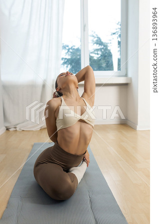 Young flexible woman practicing advanced yoga stretch indoors. Female performing deep backbend pose with leg hold on yoga mat, flexibility training, strength and balance in minimal studio. Young flexible woman practicing advanced yoga stretch indoors. Female performing deep backbend pose with leg hold on yoga mat, flexibility training, strength and balance in minimal studio. 136893964