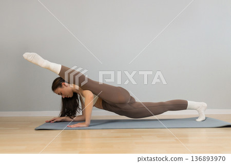 Young athletic woman performing advanced yoga plank variation indoors. Female practicing strength and flexibility exercise on yoga mat, balance control, core training, minimal studio background. Young athletic woman performing advanced yoga plank variation indoors. Female practicing strength and flexibility exercise on yoga mat, balance control, core training, minimal studio background. 136893970
