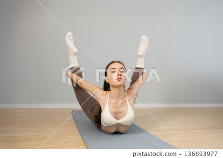 Young woman practicing advanced yoga pose indoors. Athletic female holding legs while lying on mat, strong back flexibility, balance and control, minimal studio interior Young woman practicing advanced yoga pose indoors. Athletic female holding legs while lying on mat, strong back flexibility, balance and control, minimal studio interior 136893977