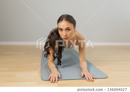 Young woman practicing yoga on mat indoors. Low plank forearm position, strong core and arms, athletic female body, focused look at camera, minimal studio interior, soft natural light. Young woman practicing yoga on mat indoors. Low plank forearm position, strong core and arms, athletic female body, focused look at camera, minimal studio interior, soft natural light. 136894027