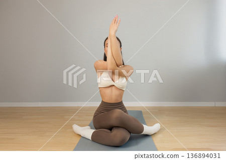Young woman practicing yoga indoors on mat. Seated twist pose with crossed legs and raised arms, flexible athletic body, calm focused mood, minimal studio interior, soft natural light. Young woman practicing yoga indoors on mat. Seated twist pose with crossed legs and raised arms, flexible athletic body, calm focused mood, minimal studio interior, soft natural light. 136894031