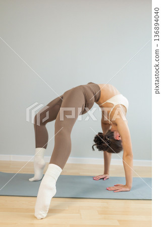 Young woman practicing advanced yoga backbend indoors. Wheel pose with strong arch, flexibility and strength training on yoga mat, minimal studio interior, wellness and mindfulness concept. Young woman practicing advanced yoga backbend indoors. Wheel pose with strong arch, flexibility and strength training on yoga mat, minimal studio interior, wellness and mindfulness concept. 136894040