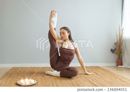 Young woman practicing advanced yoga stretching indoors. Seated pose with raised leg, flexibility and balance training, candles nearby, calm minimal interior, wellness and mindfulness concept. 136894051