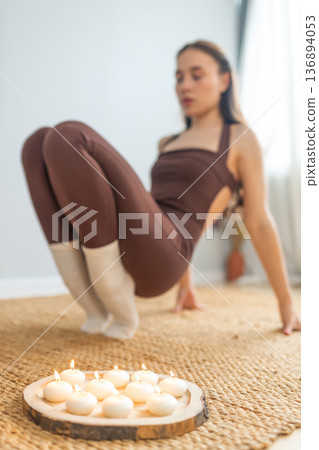 Young woman practicing yoga strength pose indoors. Balance and core training on natural mat, candles nearby, minimal calm interior, wellness, mindfulness and healthy lifestyle concept. 136894053
