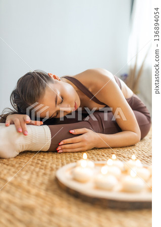 Young woman practicing yoga stretching indoors. Forward bend pose on natural mat, soft candlelight, calm minimal interior, relaxation, mindfulness and wellness lifestyle concept. Young woman practicing yoga stretching indoors. Forward bend pose on natural mat, soft candlelight, calm minimal interior, relaxation, mindfulness and wellness lifestyle concept. 136894054
