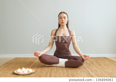 Young woman practicing meditation indoors. Lotus pose on natural mat, closed eyes, burning candles nearby, calm minimal interior, mindfulness and wellness lifestyle concept. 136894061