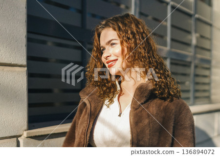 Young woman portrait near modern building facade in sunlight. Stylish casual outfit, soft shadows, confident look, contemporary urban lifestyle and fashion concept. 136894072