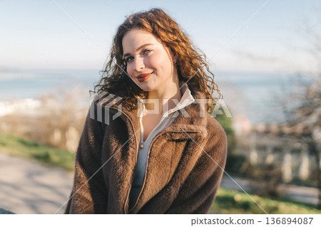 Young woman sitting on seaside promenade railing, enjoying sunny day by the sea. Casual outfit, relaxed pose, coastal city lifestyle, freedom and calm outdoor atmosphere. Young woman sitting on seaside promenade railing, enjoying sunny day by the sea. Casual outfit, relaxed pose, coastal city lifestyle, freedom and calm outdoor atmosphere. 136894087
