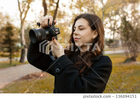 Young woman reviewing photos on her digital camera while standing in a sunny park. Warm autumn light, creative outdoor photography moment, lifestyle scene. Young woman reviewing photos on her digital camera while standing in a sunny park. Warm autumn light, creative outdoor photography moment, lifestyle scene. 136894151