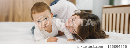 Cozy moment of a young woman and baby relaxing together at home, lying on a soft bed and smiling in natural light. Cozy moment of a young woman and baby relaxing together at home, lying on a soft bed and smiling in natural light. 136894256