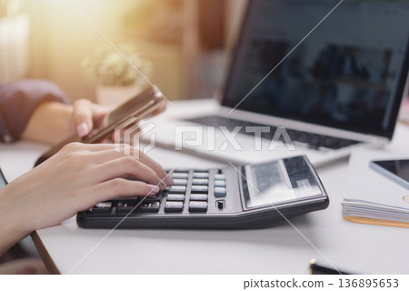 Woman hand use calculator, holding smartphone, work office desk laptop. Financial accounting concept. Calculate tax, budget, data analysis, expense report. remote work, multitasking, sunlight. Woman hand use calculator, holding smartphone, work office desk laptop. Financial accounting concept. Calculate tax, budget, data analysis, expense report. remote work, multitasking, sunlight. 136895653