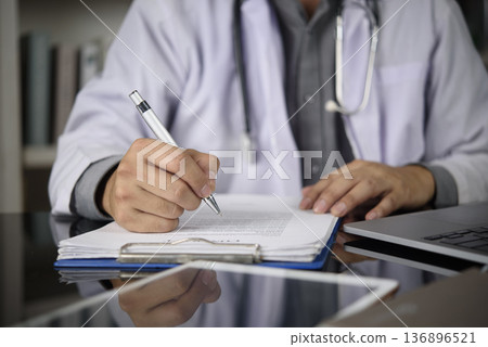 Doctor hand writing on medical clipboard, filling patient form at clinic. Physician in white coat with stethoscope writing prescription, working with laptop. Healthcare, medicine, diagnosis concept. 136896521