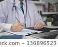 Male physician writing patient report clipboard hospital office. Professional doctor doing paperwork, signing insurance documents. Medical consultation, health record administration and job concept. 136896523