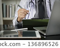 Physician workspace with doctor writing notes clipboard. Stethoscope, laptop, tablet on desk patient record management. Healthcare technology, electronic health records EHR, and consultation concept. 136896526