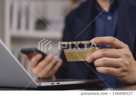 Man hand holding credit card, using smartphone for online shopping payment. Businessman making e-commerce transaction, purchasing on internet with laptop. Digital banking and secure payment concept. 136896527