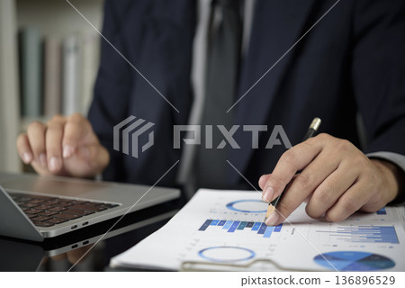 Businessman analyzing financial data report at office desk. Man hand pointing pencil at chart, graph for investment planning, audit. Business performance, market analysis, and strategy concept. Businessman analyzing financial data report at office desk. Man hand pointing pencil at chart, graph for investment planning, audit. Business performance, market analysis, and strategy concept. 136896529