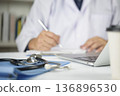 Stethoscope on desk with doctor writing patient report in background. Physician at work in hospital office, using laptop for research, telemedicine. Professional medical consultation concept. 136896530