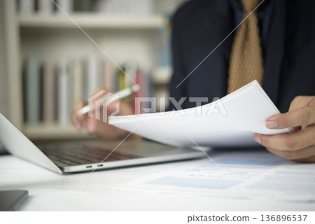 Businessman hand holding document, reviewing financial report with laptop at office desk. Manager analyzing paperwork, contract for business audit. Corporate planning and management concept. Businessman hand holding document, reviewing financial report with laptop at office desk. Manager analyzing paperwork, contract for business audit. Corporate planning and management concept. 136896537