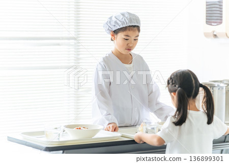 A girl on school lunch duty wearing a white lab coat and serving food from a large pot A girl on school lunch duty wearing a white lab coat and serving food from a large pot 136899301