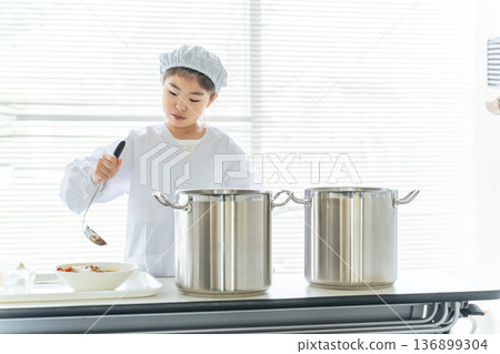 A girl on school lunch duty wearing a white lab coat and serving food from a large pot A girl on school lunch duty wearing a white lab coat and serving food from a large pot 136899304