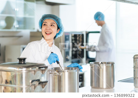 A female chef wearing a sanitary white coat cooking in a large pot in the kitchen, school lunch center, cafeteria A female chef wearing a sanitary white coat cooking in a large pot in the kitchen, school lunch center, cafeteria 136899322