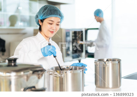 A female chef wearing a sanitary white coat cooking in a large pot in the kitchen, school lunch center, cafeteria A female chef wearing a sanitary white coat cooking in a large pot in the kitchen, school lunch center, cafeteria 136899323