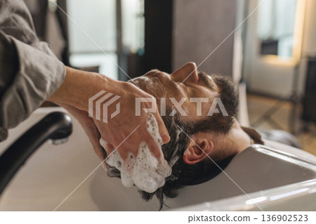 Barber washing client hair with shampoo at salon sink. Professional male grooming service, relaxing head wash process, close up of hairstyling care in modern barbershop. Barber washing client hair with shampoo at salon sink. Professional male grooming service, relaxing head wash process, close up of hairstyling care in modern barbershop. 136902523