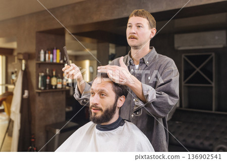 Barber preparing male client for haircut in modern barbershop. Professional barber service, beard grooming, men hairstyle care and lifestyle concept. Barber preparing male client for haircut in modern barbershop. Professional barber service, beard grooming, men hairstyle care and lifestyle concept. 136902541