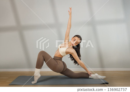 Flexible young woman practicing yoga stretch indoors. Female performing deep lunge side stretch pose on yoga mat, strength, balance and flexibility training in minimal studio with natural light. Flexible young woman practicing yoga stretch indoors. Female performing deep lunge side stretch pose on yoga mat, strength, balance and flexibility training in minimal studio with natural light. 136902737