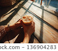 Photo of a person holding an iced latte in a cafe with sunlight filtering through the trees 136904283