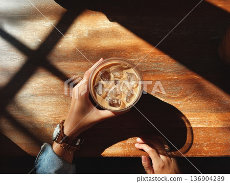 Photo of a person holding an iced latte in a cafe with sunlight filtering through the trees 136904289