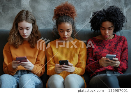 Three young women engaged in texting on smartphones while sitting closely together on a couch in a modern interior during the afternoon Three young women engaged in texting on smartphones while sitting closely together on a couch in a modern interior during the afternoon 136907001