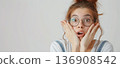 Young woman with glasses holding her cheeks in shock, her mouth open, against a plain background. 136908542