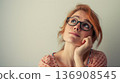Reflective young woman in glasses looks upwards, hand on chin, against a neutral background. 136908545