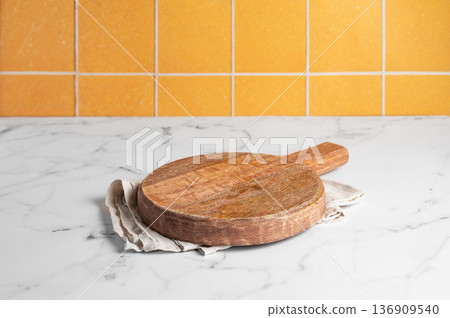 Empty wooden cutting board with a kitchen towel on a marble countertop against an orange tiled wall. Empty wooden cutting board with a kitchen towel on a marble countertop against an orange tiled wall. 136909540