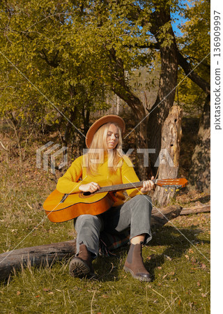 Beautiful blonde girl playing acoustic guitar and singing relaxing enjoying outdoors on sunny autumn day. Hippie woman in boho style learning and practicing playing musical instrument. Fall leaves 136909997