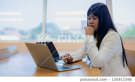 Concentrated Asian businesswoman using laptop and digital tablet with stylus pen while working in modern office, female executive pondering project strategy. 136911098
