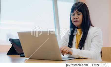 Professional Asian businesswoman focusing on laptop in bright office. Mature female entrepreneur working with digital tablet on desk. Career success, corporate lifestyle, and modern workspace. 136911099