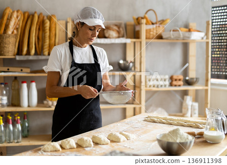 Elderly woman baker sifts flour for dough Elderly woman baker sifts flour for dough 136911398
