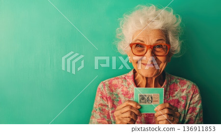 Joyful older woman holds vintage stamps against a vibrant turquoise background, showcasing her passion for collecting during a sunny afternoon at home 136911853