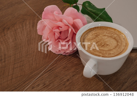 Cup of coffee rests beside a pink rose and a silver notebook on a wooden table Cup of coffee rests beside a pink rose and a silver notebook on a wooden table 136912048
