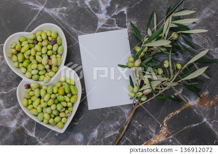 Healthy green olives in heart shaped white plate with tree branch, Mediterranean superfood rich in vitamins and minerals, blank paper for copy space 136912072