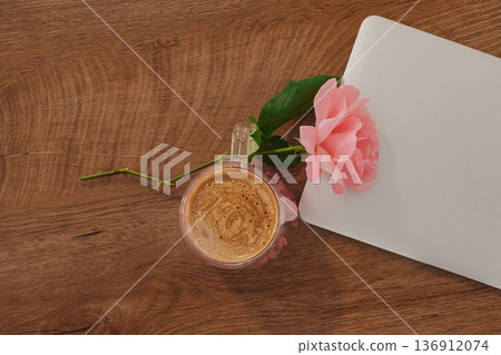 Cup of coffee rests beside a pink rose and a silver notebook on a wooden table 136912074