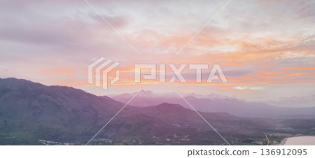 Panoramic sunset view of Crete with shadowed mountain slopes, snow-capped peaks of Lefka Ori, olive valley, coastal village lights, and pastel sky 136912095
