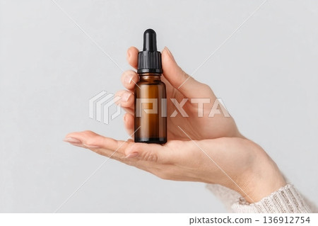 Amber glass dropper bottle in hand. Minimal skincare concept with clean white background. Cosmetic serum packaging. Elegant presentation of beauty product. Natural essential oil container. Amber glass dropper bottle in hand. Minimal skincare concept with clean white background. Cosmetic serum packaging. Elegant presentation of beauty product. Natural essential oil container. 136912754