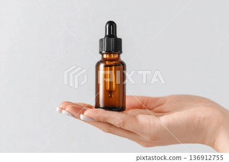 Amber glass dropper bottle in hand. Minimal skincare concept with clean white background. Cosmetic serum packaging. Elegant presentation of beauty product. Natural essential oil container. Amber glass dropper bottle in hand. Minimal skincare concept with clean white background. Cosmetic serum packaging. Elegant presentation of beauty product. Natural essential oil container. 136912755